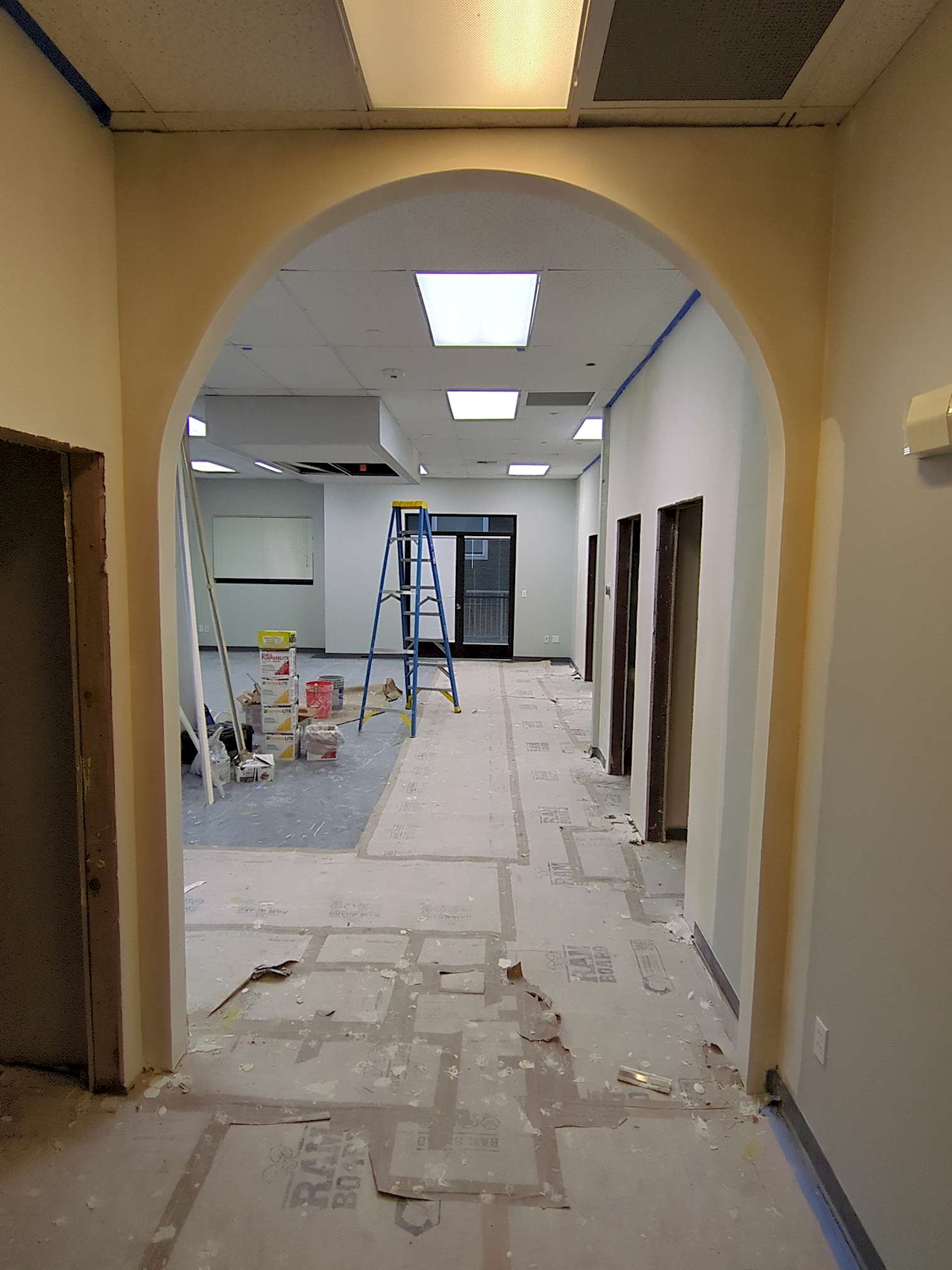 SDG construction 4 Beyond a rounded arch at the entryway sits an office space under renovation, complete with construction materials toward the left and a ladder in the center. To the sides are several empty doorways; the surrounding drywall is finished.