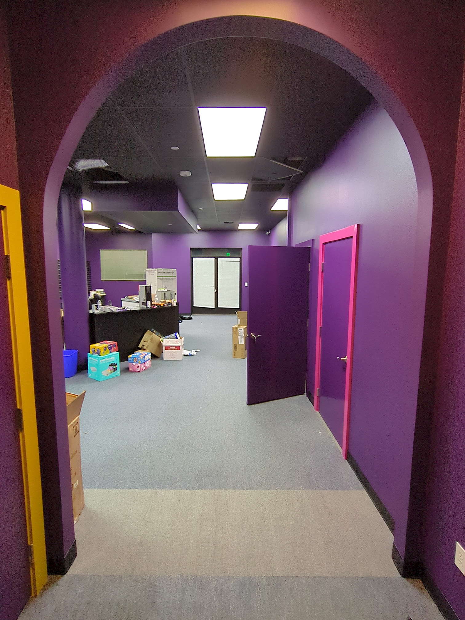 SDG construction 15 Through a tall arch one can see the now-familiar purple walls, black ceiling, and grey and slate-grey carpet of the Silver Door. Beyond and to the left, a large black counter can be seen next to a round pillar. On top of and around it are a variety of boxes, some unopened and some ready for recycling.