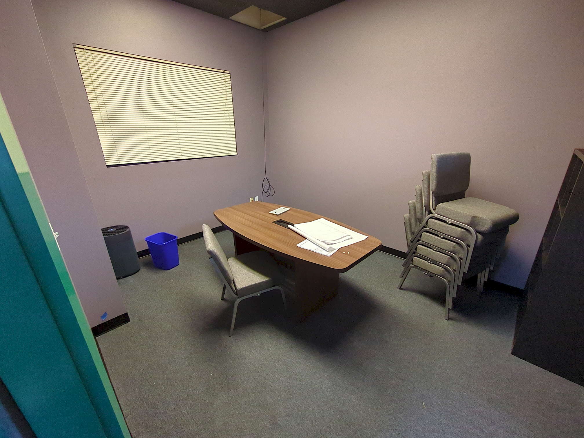 SDG construction 17 The purplish-grey interior of a game room, looking a bit bare-bones at the moment. A large air filtration unit sits in the left corner next to a small recycle bin. In the center is a grey chair in front of a long wooden table with rounded sides, on top of which rests a few large papers. To the right sits a stack of five more grey chairs and part of a medium-tall black shelving unit.